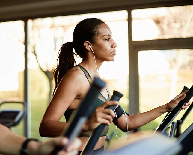 Mujer joven haciendo ejercicio en una máquina elíptica en un gimnasio. Lleva un top deportivo y auriculares.