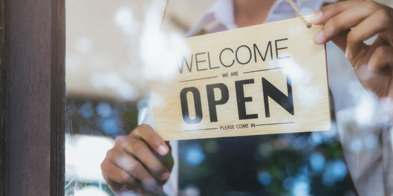 Imagen de una persona poniendo un letrero con el texto “welcome we are open please come in”