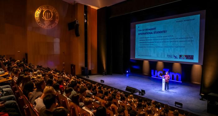 Fotografía del acto de bienvenida en el auditorio de la universidad. En el escenario un estudiante se dirige al público