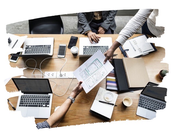 Fotografía de una mesa de trabajo con portátiles, móviles y personas cambiando documentos